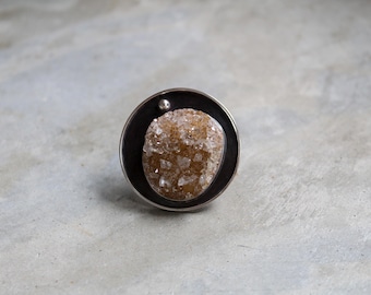 May include: A circular ring featuring a dark, oxidized metal setting that frames a textured, amber-colored druzy gemstone. A small, polished silver bead is positioned at the top edge of the circular frame, contrasting with the dark metal.
