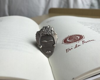 May include: A silver-toned metal ring shaped like a stylized face with detailed hair, prominent eyebrows, and small circular accents at the jawline. The ring rests on an open book page featuring a circular red stamp and the handwritten text Fri da Rivera.