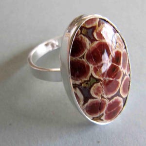 Puede incluir: Anillo de plata con una piedra ovalada. La piedra presenta un patrón de manchas granate y crema. La banda del anillo es de plata pulida lisa.