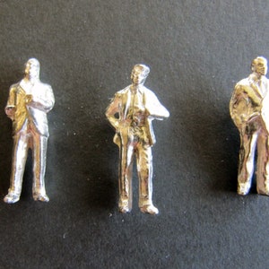 Op de afbeelding: Drie zilveren miniatuurfiguurtjes van mensen in pak. De figuurtjes staan rechtop en zijn ongeveer 5 cm hoog.