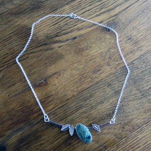 Puede incluir: Collar de plata con una piedra verde ovalada con manchas oscuras. La cadena fina se complementa con elementos en forma de hojas a cada lado de la piedra, creando un dise&ntilde;o inspirado en la naturaleza.