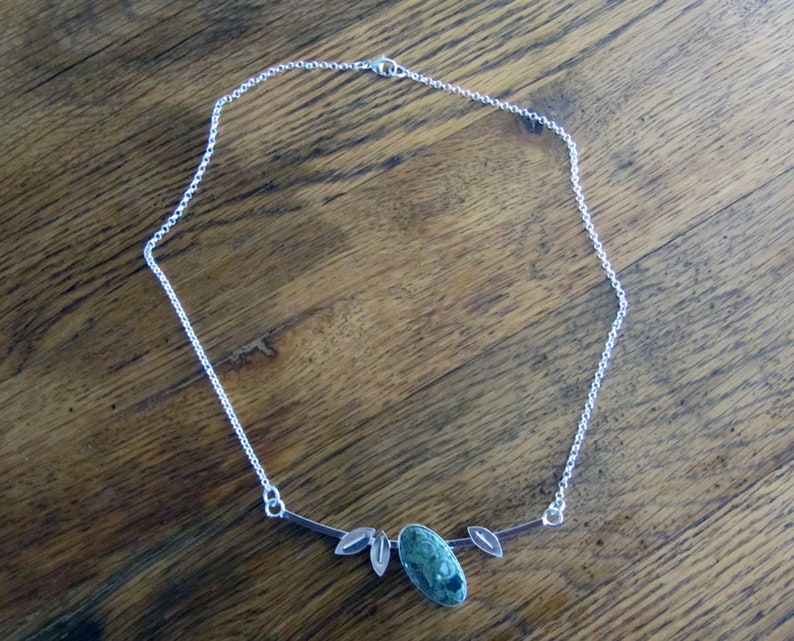Puede incluir: Collar de plata con una piedra verde ovalada con manchas oscuras. La cadena fina se complementa con elementos en forma de hojas a cada lado de la piedra, creando un dise&ntilde;o inspirado en la naturaleza.