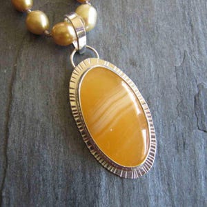 May include: A necklace featuring a large, oval-shaped, golden-yellow banded agate pendant set in a silver bezel with a textured, notched edge. The pendant hangs from a strand of round, lustrous, golden-toned pearls that are strung together.