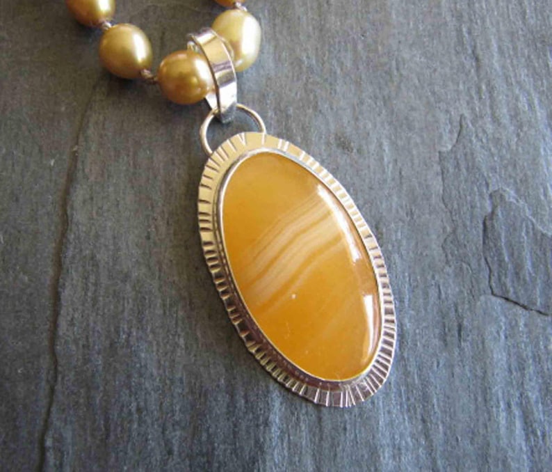 May include: A necklace featuring a large, oval-shaped, golden-yellow banded agate pendant set in a silver bezel with a textured, notched edge. The pendant hangs from a strand of round, lustrous, golden-toned pearls that are strung together.