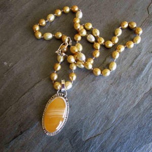 May include: A necklace featuring a strand of golden-hued pearls and a large, oval-shaped yellow agate pendant. The pendant is encased in a textured silver-toned metal setting with a notched edge, connected to the pearl strand by a silver-toned bail.