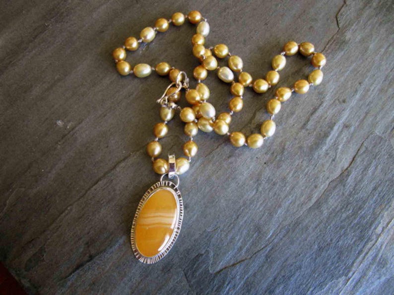 May include: A necklace featuring a strand of golden-hued pearls and a large, oval-shaped yellow agate pendant. The pendant is encased in a textured silver-toned metal setting with a notched edge, connected to the pearl strand by a silver-toned bail.