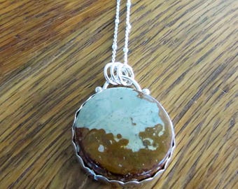 May include: A round pendant necklace with a silver chain. The pendant features a stone with a mix of brown and light green colors, set in a silver bezel. The necklace is displayed on a wooden surface.