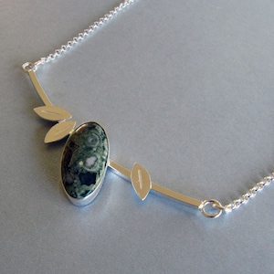 Puede incluir: Un collar de plata con un colgante ovalado de piedra verde y negra. El colgante est&aacute; adornado con detalles plateados en forma de hoja y una fina cadena. El collar es una pieza inspirada en la naturaleza.