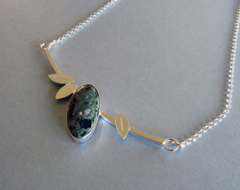 Puede incluir: Un collar de plata con un colgante ovalado de piedra verde y negra. El colgante est&aacute; adornado con detalles plateados en forma de hoja y una fina cadena. El collar es una pieza inspirada en la naturaleza.