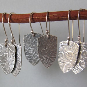 May include: Five pairs of silver dangle earrings with a floral pattern. The earrings are hanging from a brown branch.
