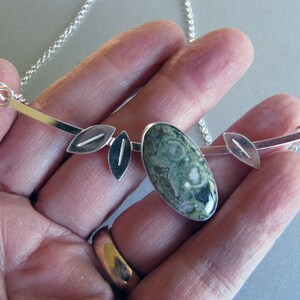 Puede incluir: Collar de plata con un colgante ovalado de piedra verde moteada. El colgante se complementa con detalles plateados en forma de hoja y una fina cadena. Este collar de inspiraci&oacute;n natural es adecuado para el uso diario.