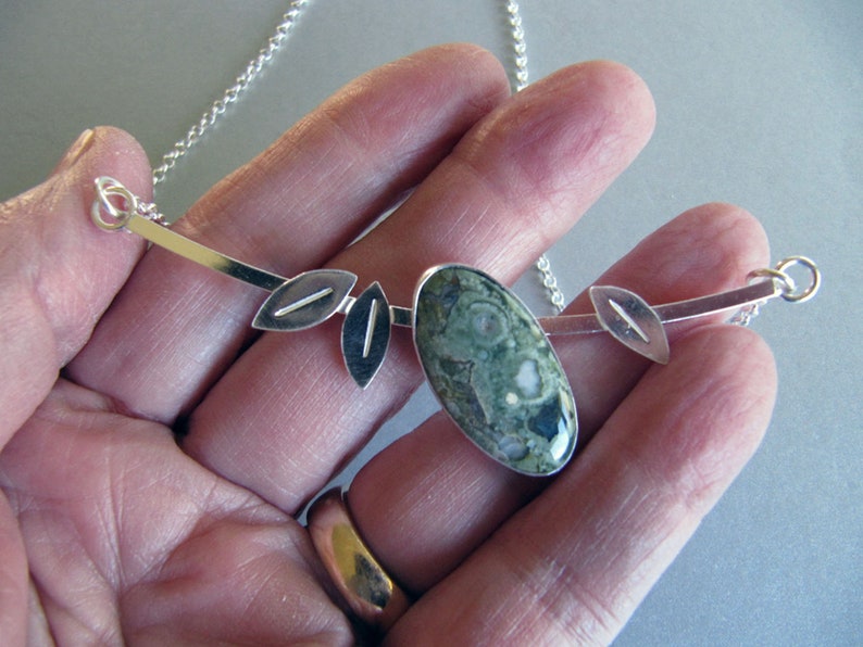 Puede incluir: Collar de plata con un colgante ovalado de piedra verde moteada. El colgante se complementa con detalles plateados en forma de hoja y una fina cadena. Este collar de inspiraci&oacute;n natural es adecuado para el uso diario.