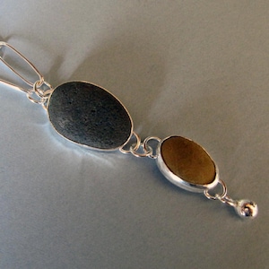 May include: A silver necklace featuring two oval-shaped stones. One stone is dark gray, the other is brown. The stones are set in silver bezels and connected by a silver chain. A small silver ball hangs at the end.