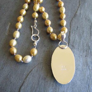 May include: A necklace featuring a strand of golden-hued pearls with a silver hook clasp. Attached is an oval-shaped silver pendant with a smooth, flat back surface. The metal surface of the pendant is engraved with the letters KF and the word STERLING.