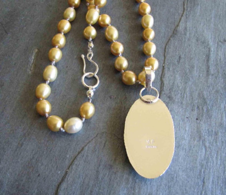 May include: A necklace featuring a strand of golden-hued pearls with a silver hook clasp. Attached is an oval-shaped silver pendant with a smooth, flat back surface. The metal surface of the pendant is engraved with the letters KF and the word STERLING.
