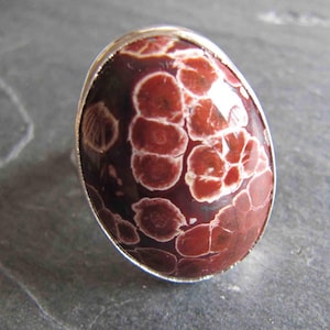 Puede incluir: Anillo ovalado con una piedra grande y pulida. La piedra tiene una base granate con un patrón de formas rojas y blancas. Está engastada en una banda de metal plateado. Una joya distintiva y elegante.