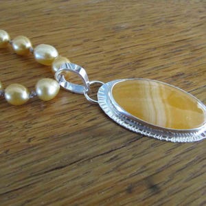 May include: A necklace featuring a strand of lustrous golden pearls connected to a large oval pendant. The pendant consists of a smooth, banded yellow stone set in a polished silver frame with a decorative, textured edge and a silver bail.
