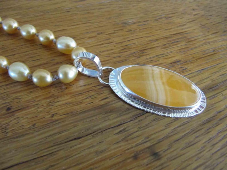 May include: A necklace featuring a strand of lustrous golden pearls connected to a large oval pendant. The pendant consists of a smooth, banded yellow stone set in a polished silver frame with a decorative, textured edge and a silver bail.
