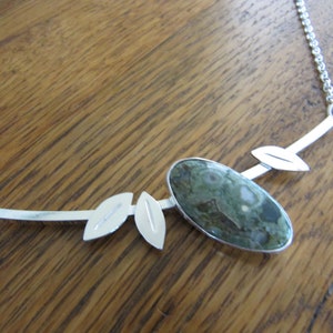 Puede incluir: Collar de plata con una piedra central ovalada verde y blanca. El collar tiene un dise&ntilde;o delicado con detalles de hojas plateadas y una cadena.
