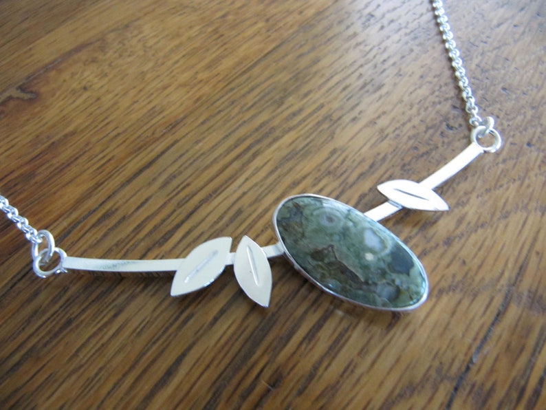 Puede incluir: Collar de plata con una piedra central ovalada verde y blanca. El collar tiene un dise&ntilde;o delicado con detalles de hojas plateadas y una cadena.