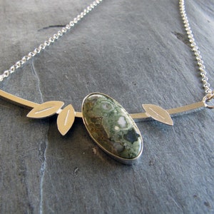 Puede incluir: Collar de plata con un colgante ovalado de piedra verde y blanca. El colgante est&aacute; engastado en plata y tiene detalles en forma de hojas. La cadena es fina.