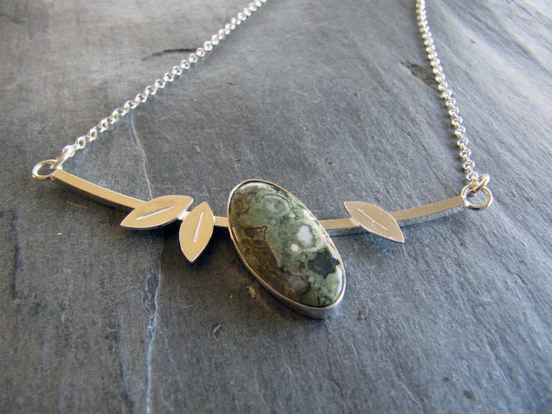 Puede incluir: Collar de plata con un colgante ovalado de piedra verde y blanca. El colgante est&aacute; engastado en plata y tiene detalles en forma de hojas. La cadena es fina.