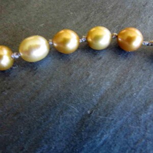 May include: A strand of lustrous, golden-hued pearls arranged in a horizontal line. Each individual pearl is round and exhibits a smooth, reflective surface. The pearls are connected by a thin, light-colored thread knotted between each bead.