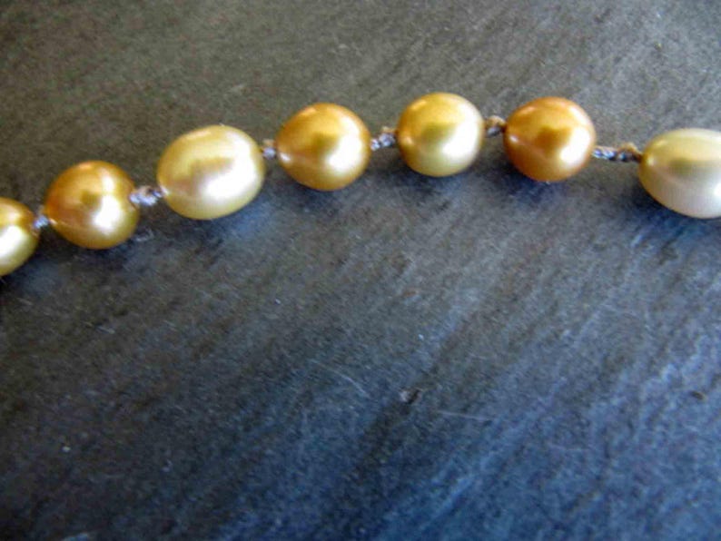 May include: A strand of lustrous, golden-hued pearls arranged in a horizontal line. Each individual pearl is round and exhibits a smooth, reflective surface. The pearls are connected by a thin, light-colored thread knotted between each bead.