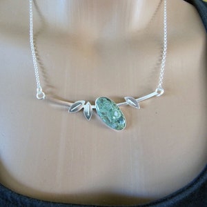 Puede incluir: Collar de plata con un colgante de piedra verde ovalada, de aproximadamente 2,5 cm de largo, con un patr&oacute;n distintivo. El colgante est&aacute; engastado en plata y tiene detalles en forma de hoja. El collar tiene una fina cadena de plata.
