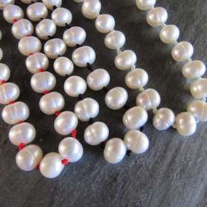 Puede incluir: Una hebra de perlas blancas enhebradas en un cordón rojo, con una segunda hebra de perlas enhebradas en un cordón blanco. Las perlas son redondas y lisas, y los cordones están anudados entre cada perla.