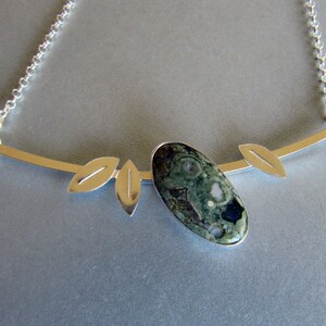 Puede incluir: Collar plateado con un colgante ovalado de piedra verde y negra. El colgante est&aacute; adornado con tres detalles en forma de hoja y una fina cadena. Una joya inspirada en la naturaleza.
