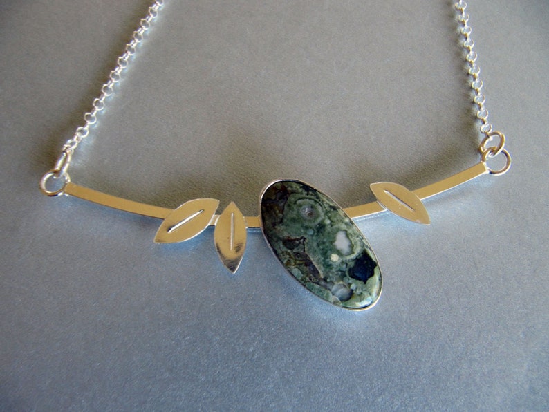 Puede incluir: Collar plateado con un colgante ovalado de piedra verde y negra. El colgante est&aacute; adornado con tres detalles en forma de hoja y una fina cadena. Una joya inspirada en la naturaleza.