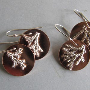 May include: Two pairs of circular copper earrings featuring a raised, textured silver-toned branch or coral-like motif on the front. Each earring is attached to a simple silver-toned metal hook for hanging, creating a contrasting metallic design.