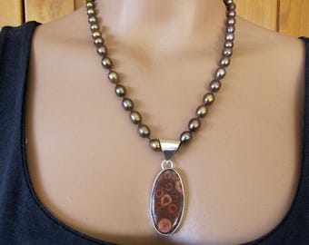May include: A necklace with a strand of dark, olive-green pearls. A silver pendant with an oval, reddish-brown stone with circular patterns hangs from the necklace. The pendant is approximately 5 cm long.
