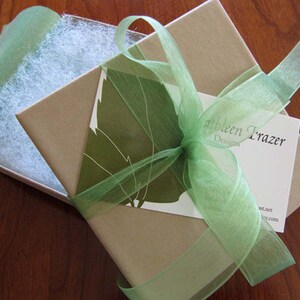 Puede incluir: Una caja de regalo envuelta en papel marr&oacute;n claro y atada con una cinta verde claro transparente. La caja est&aacute; abierta, revelando un lecho de relleno blanco. Una tarjeta de visita con el nombre "Kathleen Frazer Designs" est&aacute; metida debajo de la cinta.