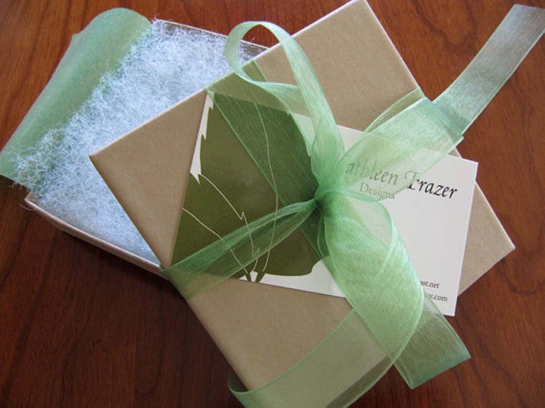Puede incluir: Una caja de regalo envuelta en papel marr&oacute;n claro y atada con una cinta verde claro transparente. La caja est&aacute; abierta, revelando un lecho de relleno blanco. Una tarjeta de visita con el nombre "Kathleen Frazer Designs" est&aacute; metida debajo de la cinta.
