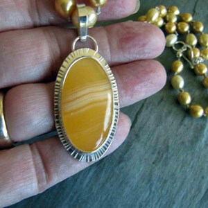May include: An oval pendant featuring a smooth, banded yellow stone set in a textured silver frame, suspended from a necklace of round, golden-hued pearls. The pendant has a polished finish with visible horizontal layers in the stone's center.
