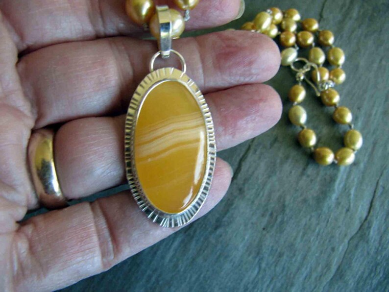 May include: An oval pendant featuring a smooth, banded yellow stone set in a textured silver frame, suspended from a necklace of round, golden-hued pearls. The pendant has a polished finish with visible horizontal layers in the stone's center.