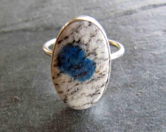 Peut inclure: Bague ovale avec une pierre blanche et grise ornée d'un centre bleu vif. La pierre est sertie dans un anneau argenté. Le design de la bague est simple mais attrayant, adapté au quotidien ou aux occasions spéciales.