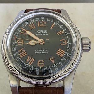 May include: An Oris automatic wristwatch with a dark grey textured dial, copper-colored Roman numerals, and a date pointer hand. The watch features a silver-toned fluted bezel, a brown leather strap, and the text Oris 25 Jewels Automatic Swiss Made.