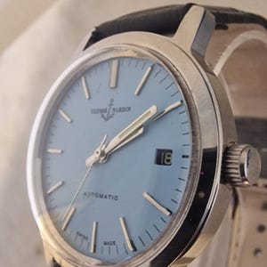 May include: A vintage Ulysse Nardin automatic wristwatch with a light blue face and silver hands and markers. The watch has a date display at the 3 o'clock position and a black leather band. The watch is marked "Swiss Made".