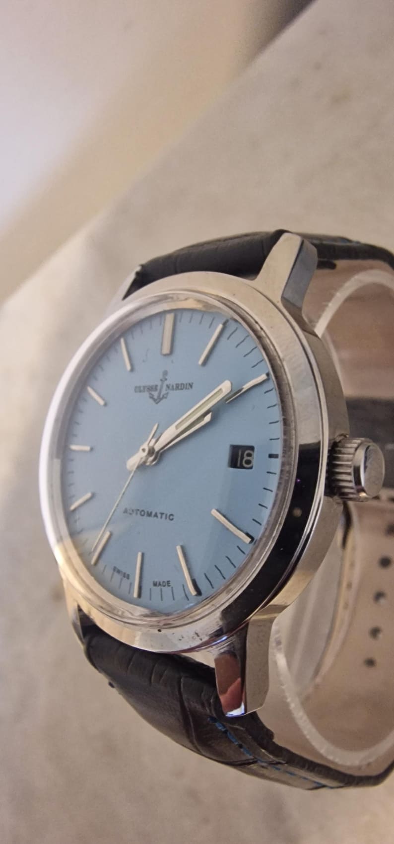 May include: A vintage Ulysse Nardin automatic wristwatch with a light blue face and silver hands and markers. The watch has a date display at the 3 o'clock position and a black leather band. The watch is marked "Swiss Made".
