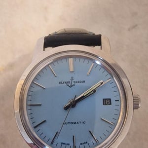 May include: A silver-toned Ulysse Nardin automatic wristwatch with a light blue face and black leather band. The watch face displays the date, hour markers, and the brand name. The watch is marked "Swiss Made".