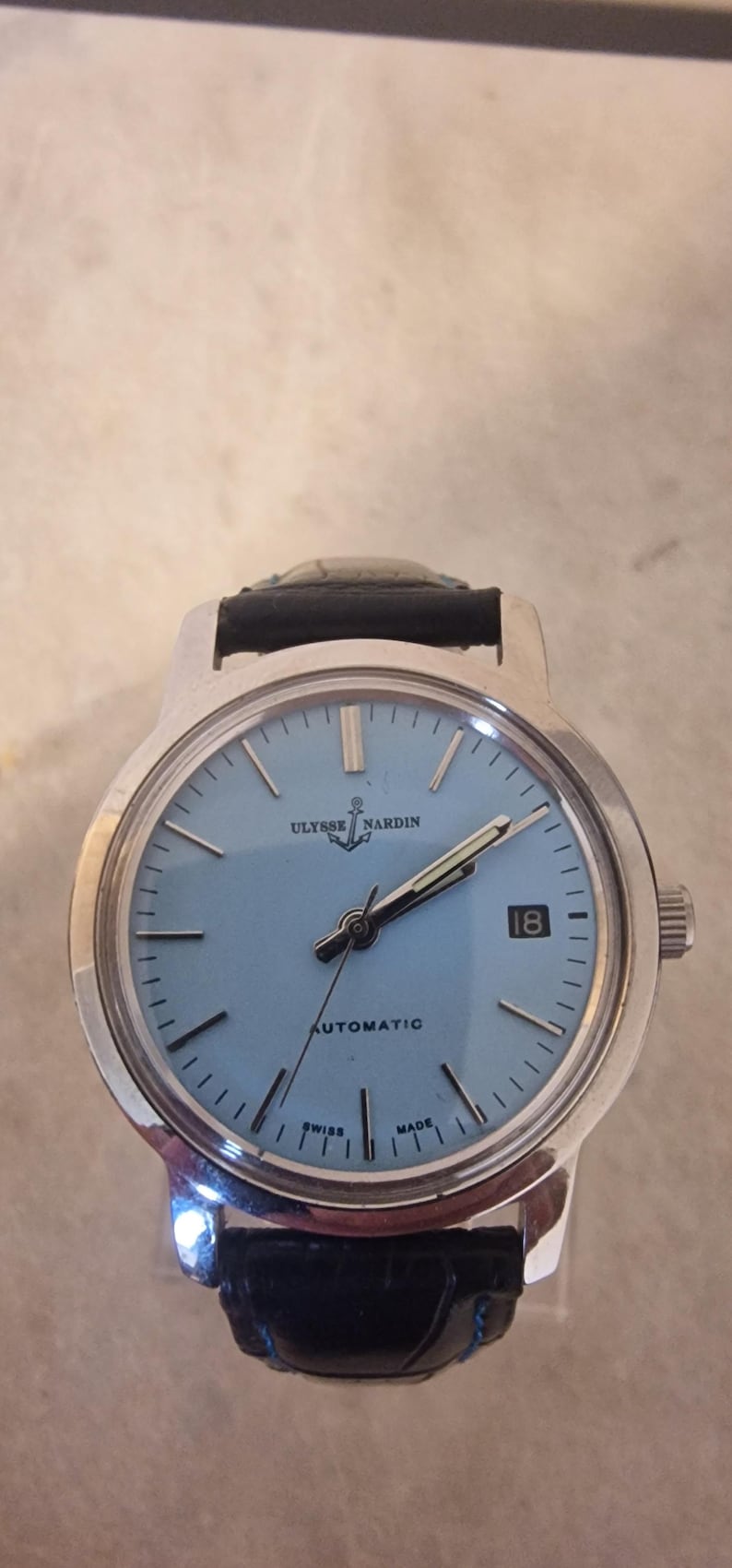 May include: A silver-toned Ulysse Nardin automatic wristwatch with a light blue face and black leather band. The watch face displays the date, hour markers, and the brand name. The watch is marked "Swiss Made".