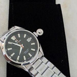 Könnte beinhalten: Eine silberne Armbanduhr mit schwarzem Zifferblatt und Datumsanzeige auf 3 Uhr. Die Uhr hat ein Edelstahlarmband und liegt auf einem schwarzen Samtbeutel.