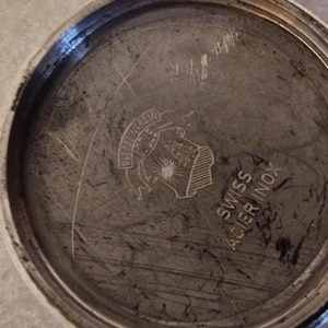 May include: Close-up of a watch back with a circular, brushed metal finish. The watch back has a stamped emblem and the text "SWISS ACIER INOX". The metal has scratches and wear.