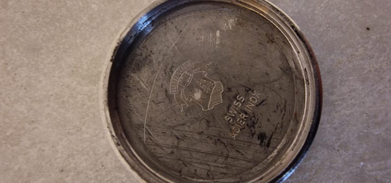 May include: Close-up of a watch back with a circular, brushed metal finish. The watch back has a stamped emblem and the text "SWISS ACIER INOX". The metal has scratches and wear.
