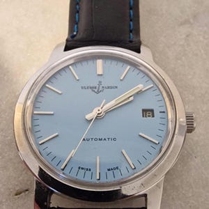 May include: A Ulysse Nardin automatic wristwatch with a light blue face and silver hands. The watch has a black leather band with blue stitching. The watch face displays the date and the words "AUTOMATIC", "SWISS", and "MADE."