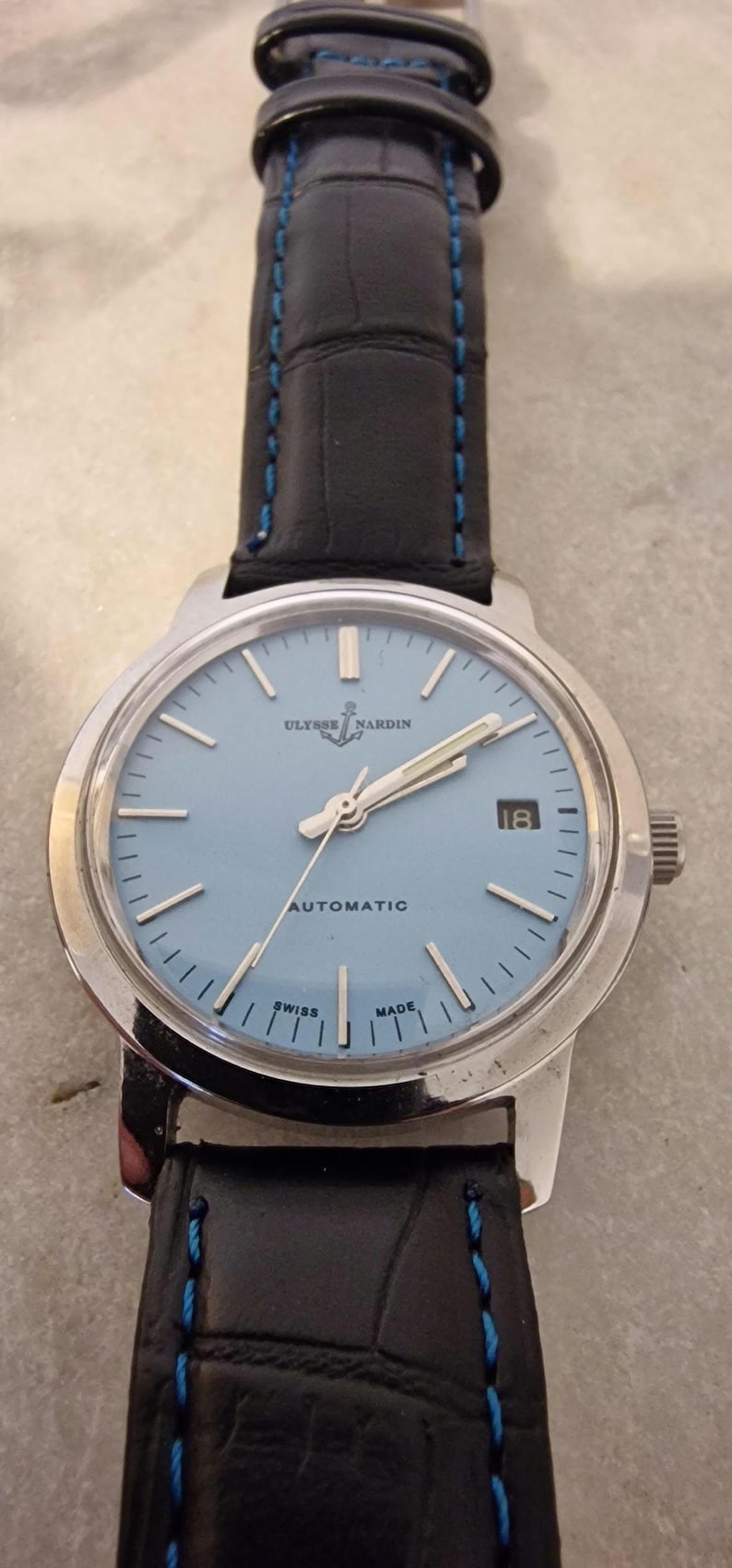 May include: A Ulysse Nardin automatic wristwatch with a light blue face and silver hands. The watch has a black leather band with blue stitching. The watch face displays the date and the words "AUTOMATIC", "SWISS", and "MADE."