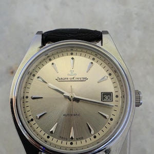 May include: A silver Jaeger-LeCoultre automatic watch with a black leather strap. The watch face is a light champagne colour with silver hour markers and hands. The date is displayed at the 3 o'clock position. The watch is marked "Swiss Made".